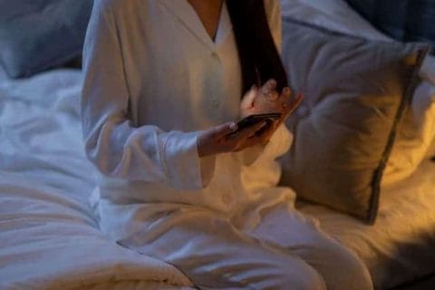Frau im Pyjama mit Smartphone in der Hand sitzt auf dem Bett