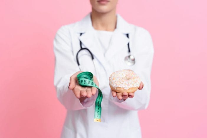 Ein Arzt hält einen Krapfen mit Zuckerguss in der einen und einen Zentimeter in der anderen Hand