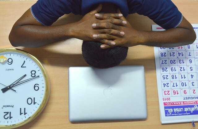  Ein übermüdeter Mann legt seinen Kopf auf den Schreibtisch, neben seinem Laptop, seinem Kalender und seiner Uhr
